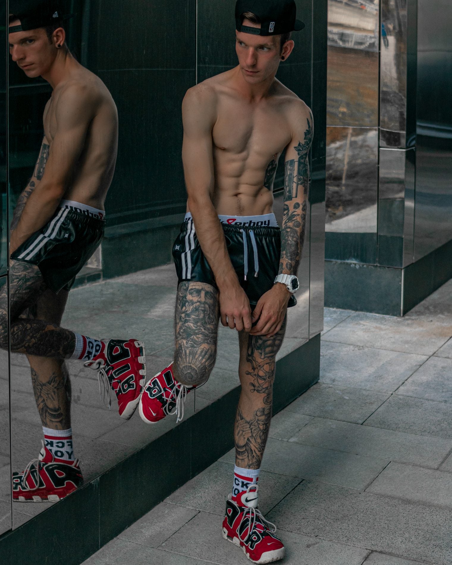sportlicher gay vor einem spiegel mit boxer berlin short in schwarz traegt fck you socks von sk8erboy socken mit roten nike air sneaker und zeigt seine tattoos