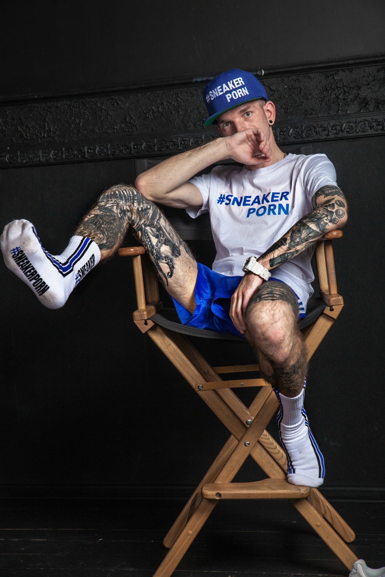 cooler junge in einem regie stuhl mit weiss blauem t-shirt und sneakerporn socken von sk8erboy zeigt seine tattoos und die sohle seiner socken mit der großen schirft