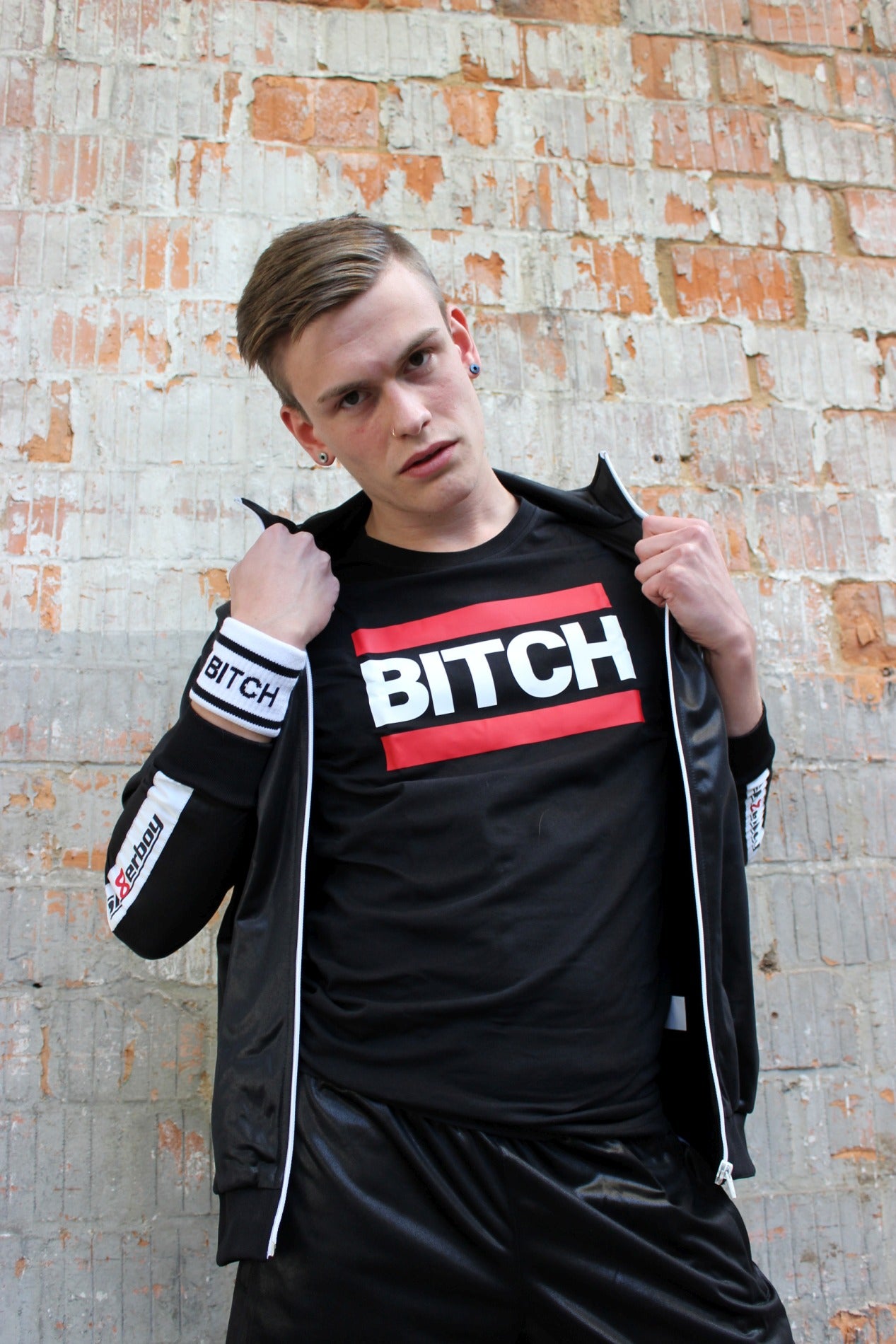 junger gay mit shiny tracksuite jacke offen zeigt ein schwarzes t-shirt mit dem aufdruck bitch in weiss und roten balken drueber und drunter